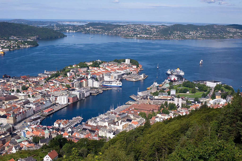 Fotografi av Bergen sett fra Fløien. Flott utsikt over byens havn og husene på hver side, sjøen og lave fjell i det fjerne under en blå himmel.