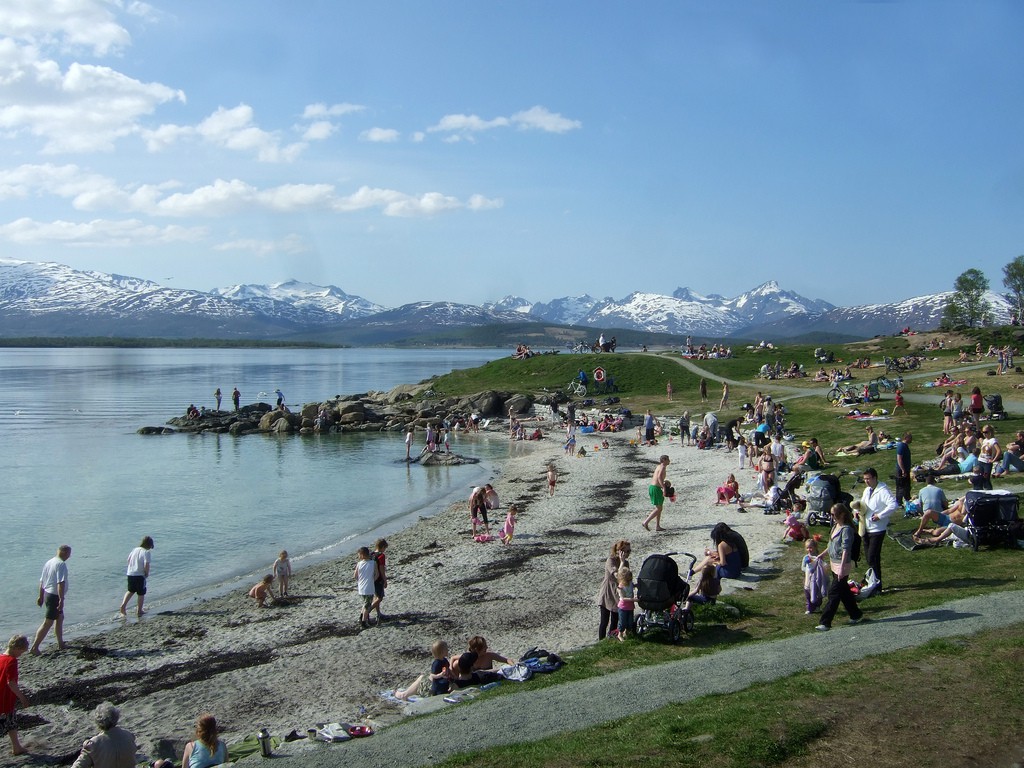 Panoramafoto av stranda Telegrafbukta i Tromsø. Mange folk er på stranden og leker ved vannkanten, og i horisonten ser vi både vann og de flotte fjellene i Tromsø under en blå himmel med noen få, avlange skyer på.